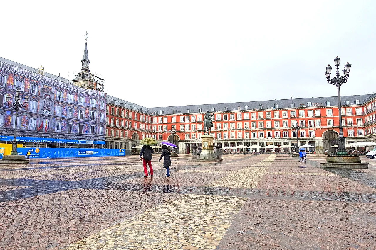 Plaza Mayor in Madrid on a rainy day, with its red-ochre façades, the equestrian statue and people with umbrellas